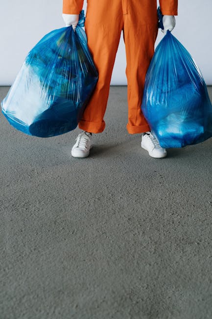 Three large black and white plastic rubbish bags filled with waste are stacked on the pavement next to a black metal fence, with a row of lush, leafy greenery behind it. The bags appear to be torn or 