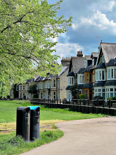 A paved pathway in a residential area with two black waste bins positioned on a grassy verge in the foreground. Behind the bins, there is a black wooden fence that runs parallel to a row of traditiona